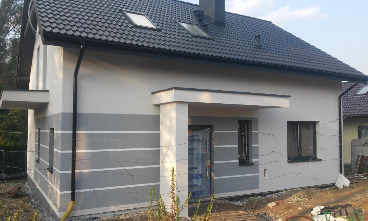 Nowoczesny dom jednorodzinny z szarą elewacją, białymi pasami dekoracyjnymi i ciemną dachówką, widoczne okna dachowe i częściowo zakryte wejście folią ochronną.