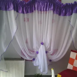 Delikatna, biała firanka z fioletowymi akcentami i dekoracyjnymi kwiatkami, upięta w kokony. W tle widoczny grzejnik i krzesło z poduszką w kratę.
