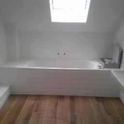 Biała wanna obudowana płytkami typu metro, umywalka i drewnopodobna podłoga w jasnej łazience na poddaszu z oknem dachowym.