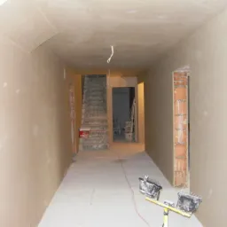 Wnętrze korytarza w trakcie wykańczania, ściany pokryte jasną warstwą, widoczne ślady po szpachlowaniu, na podłodze reflektory budowlane, w tle schody i otwory drzwiowe z odsłoniętą cegłą.