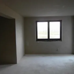 Surowe wnętrze pokoju z oknem i drzwiami balkonowymi, ściany pokryte jasnym tynkiem przed dalszą obróbką, widoczne przewody elektryczne.