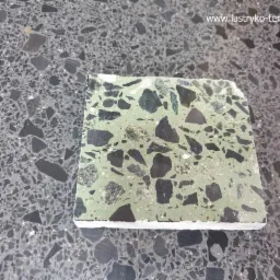 Próbka szlifowanego lastryko w odcieniach zieleni i czerni, leżąca na tle ciemnoszarej posadzki lastryko o drobniejszym ziarnie.