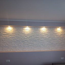 EBIMONT Sebastian Bogdanowicz - Ściana z białych, dekoracyjnych paneli imitujących kamień, oświetlona czterema punktowymi lampkami halogenowymi, zamontowana w salonie z szarymi ścianami.