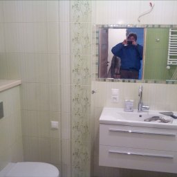 EBIMONT Sebastian Bogdanowicz - Wykończona łazienka z beżowymi płytkami z motywem roślinnym, białą szafką pod umywalką i lustrem, w którym odbija się osoba robiąca zdjęcie. Na umywalce leżą narzędzia glazurnicze.