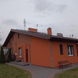 EBIMONT Sebastian Bogdanowicz - Dwukondygnacyjny dom z pomarańczową elewacją, ceglanymi elementami dekoracyjnymi i brązowym dachem, widoczny z perspektywy ogrodu z ławką i ścieżką.