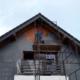 EBIMONT Sebastian Bogdanowicz - Ocieplanie elewacji budynku jednorodzinnego z użyciem rusztowania, widoczna warstwa izolacyjna z szarego styropianu oraz fragment ściany z czerwonej cegły.