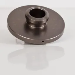 Precyzyjnie wykonany, metalowy element w kształcie dysku z centralnym otworem, widoczny mały otwór boczny, leżący na białej, odbijającej światło powierzchni.