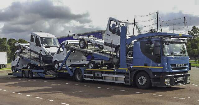 Niebieska laweta Iveco przewozi kilka białych samochodów dostawczych na platformach, widok z boku na parkingu, pochmurne niebo w tle.