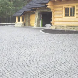 Szara kostka granitowa ułożona przed drewnianym domem z garażem. Widoczne elementy wykończeniowe i rabata. Całość tworzy estetyczny wygląd posesji.