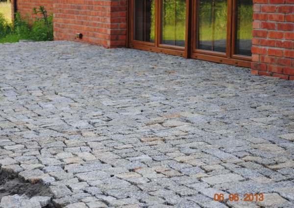 Ułożona szara kostka granitowa na podwórku przy ceglanym budynku z oknami. Widoczny fragment trawnika. Data na dole zdjęcia.