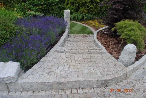 Granitowa ścieżka z delikatnymi schodkami, otoczona bujną roślinnością, w tym fioletową lawendą, prowadząca przez trawnik i rabatę z korą.