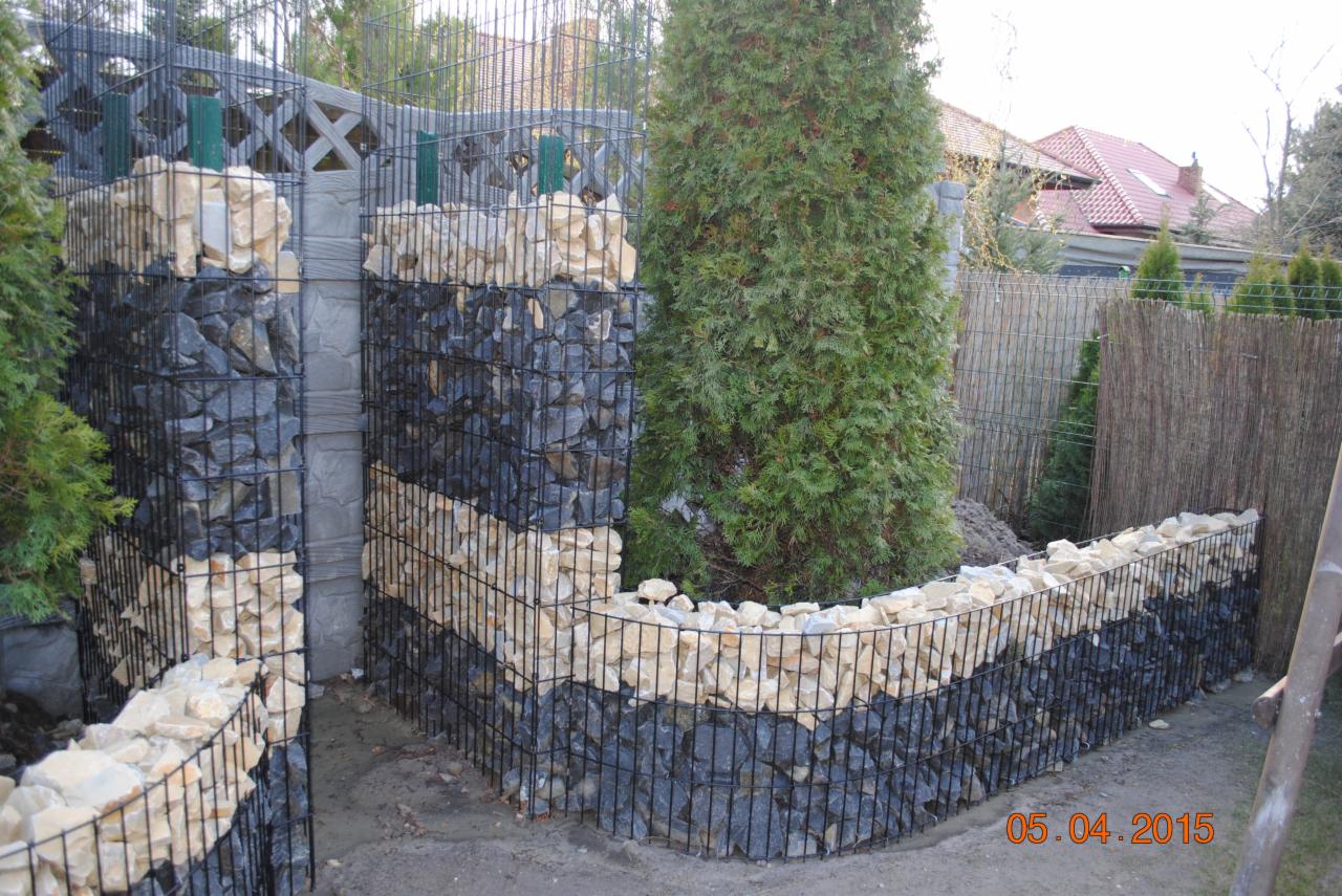 Gabionowe ogrodzenie z kamienia łupanego, ułożonego w pasy. Konstrukcja z czarnej siatki otacza drzewo iglaste. W tle widoczny dom z czerwoną dachówką i ogrodzenie z trzciny.