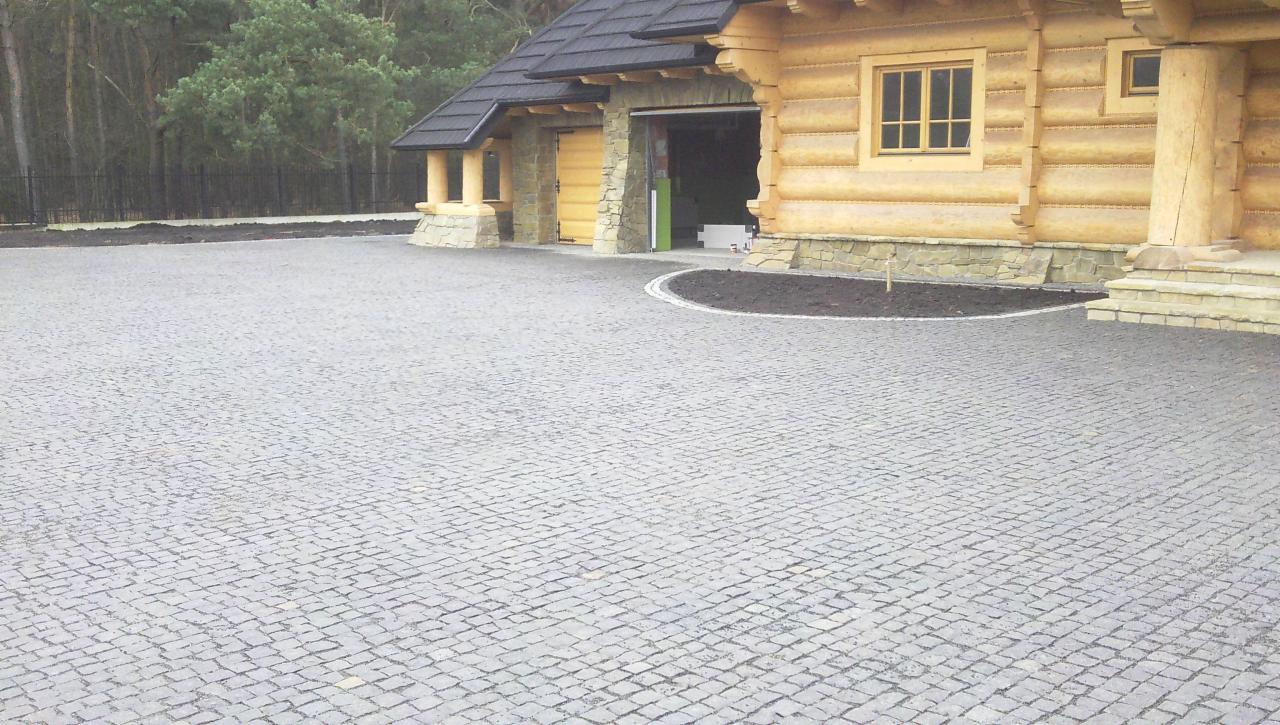 Szara kostka granitowa ułożona przed drewnianym domem z garażem. Widoczne elementy wykończeniowe i rabata. Całość tworzy estetyczny wygląd posesji.