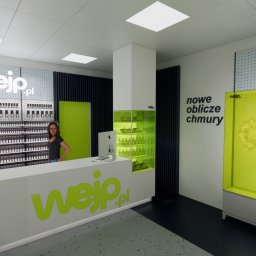 Projektownia - Nowoczesne wnętrze biura z ladą recepcyjną w kolorze białym i zielonym, regałami z produktami oraz kobietą stojącą za ladą. Oświetlenie sufitowe, minimalistyczny design.