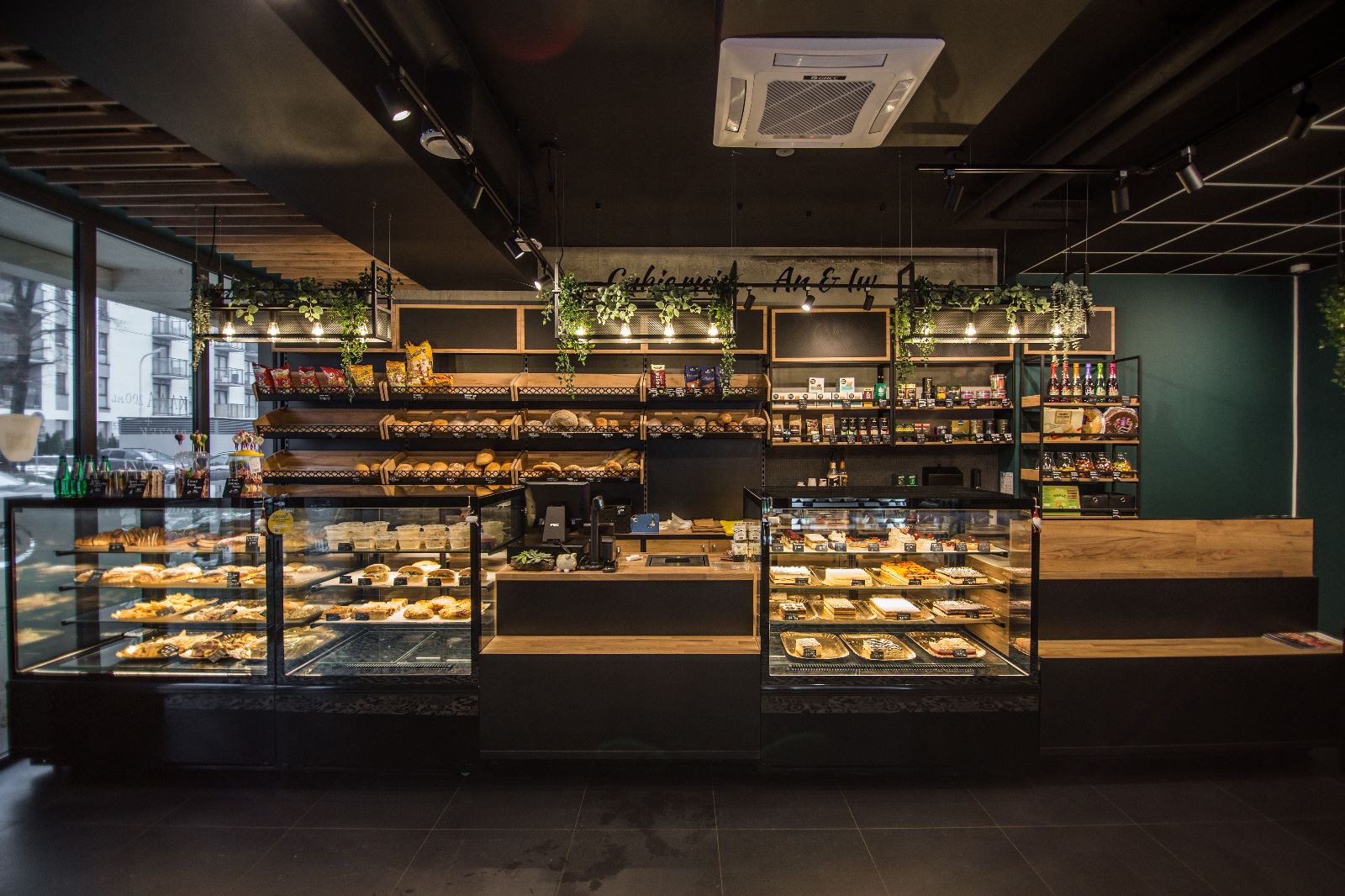 Nowoczesny wystrój cukierni: ciemne ściany, drewniane regały z pieczywem i słodyczami, przeszklone witryny z ciastami, wiszące rośliny i oświetlenie punktowe.
