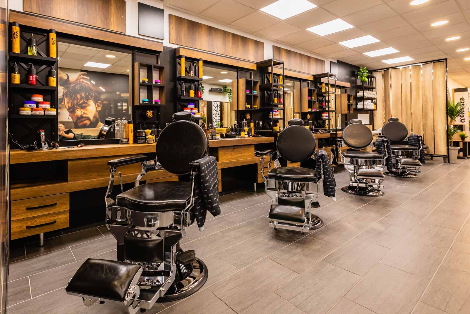 Nowoczesne wnętrze barber shopu z czterema skórzanymi fotelami fryzjerskimi, drewnianymi elementami wystroju i lustrami z kosmetykami. Stonowana kolorystyka.