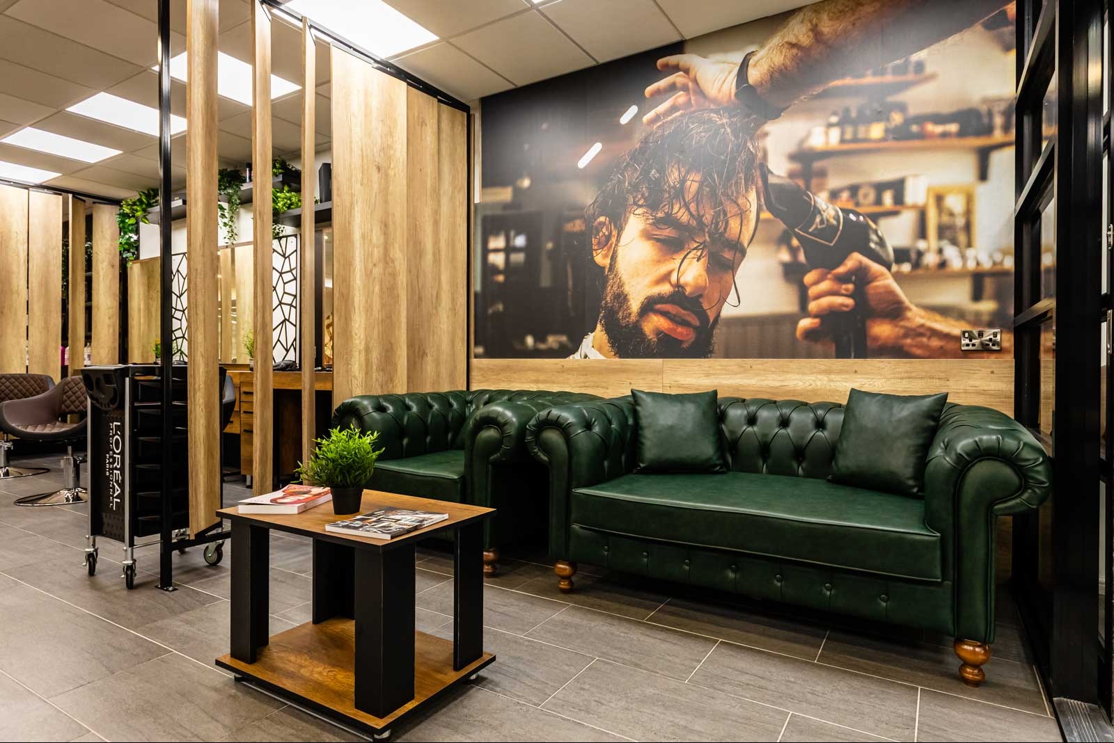 Stylowa poczekalnia w salonie fryzjerskim: zielona sofa Chesterfield, stolik kawowy z książkami, ściana z muralem fryzjerskim i elementami drewna.