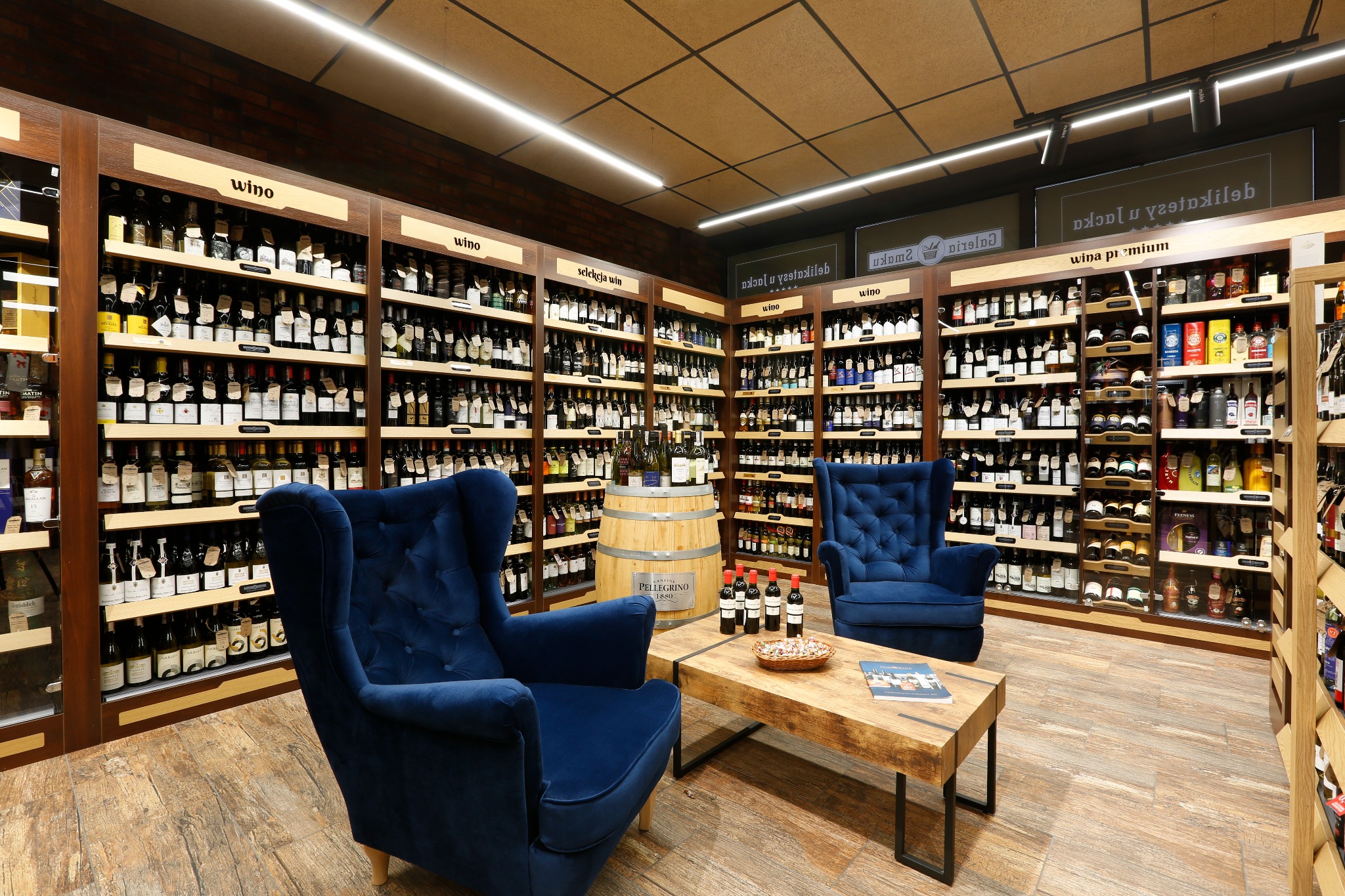 Ekskluzywny sklep z winami: ciemne drewniane regały wypełnione butelkami, dwa granatowe fotele klubowe i rustykalny stolik kawowy z przekąskami.