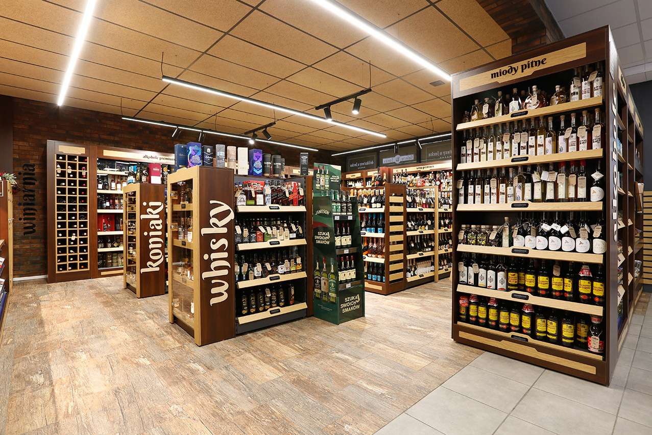 Wnętrze sklepu z alkoholem: drewniane regały z koniakami, whisky i miodami pitnymi. Ciepłe oświetlenie, brązowa cegła, podłoga imitująca drewno. Widok na alejki między regałami.