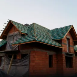 Surowy budynek z czerwonej cegły w trakcie budowy dachu z zielonej dachówki, widoczne zabezpieczenia folią okien i otworów. W tle krajobraz z górami o zmierzchu.