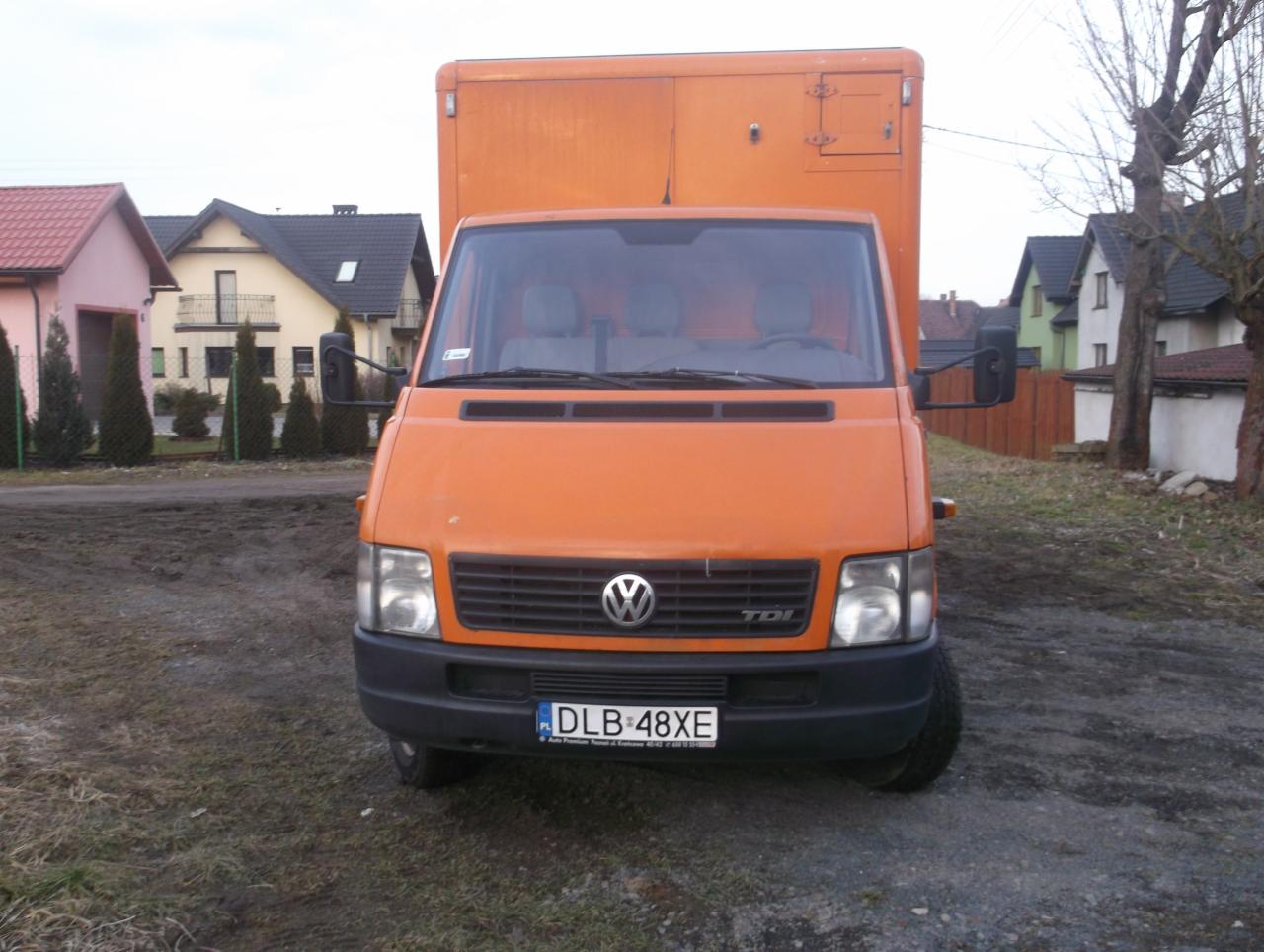 Pomarańczowy bus Volkswagen z zabudową kontenerową na tle domów w Lubaniu. Widok z przodu, tablica rejestracyjna DLB-48XE.