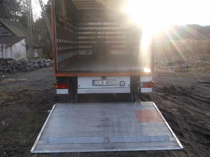 Otwarta paka busa z rampą załadunkową, widziana od tyłu, z widoczną tablicą rejestracyjną DLB 48XE, na błotnistym podłożu i w silnym oświetleniu słonecznym.