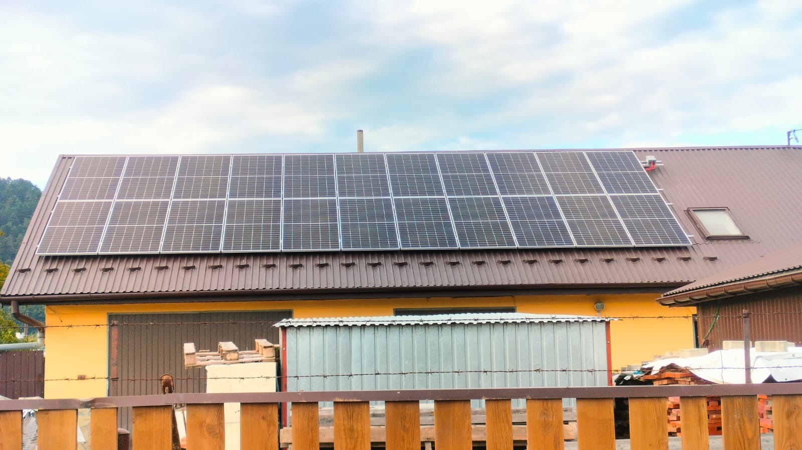 Panele słoneczne na brązowym dachu żółtego budynku gospodarczego z widoczną drewnianą palisadą i stertą cegieł.