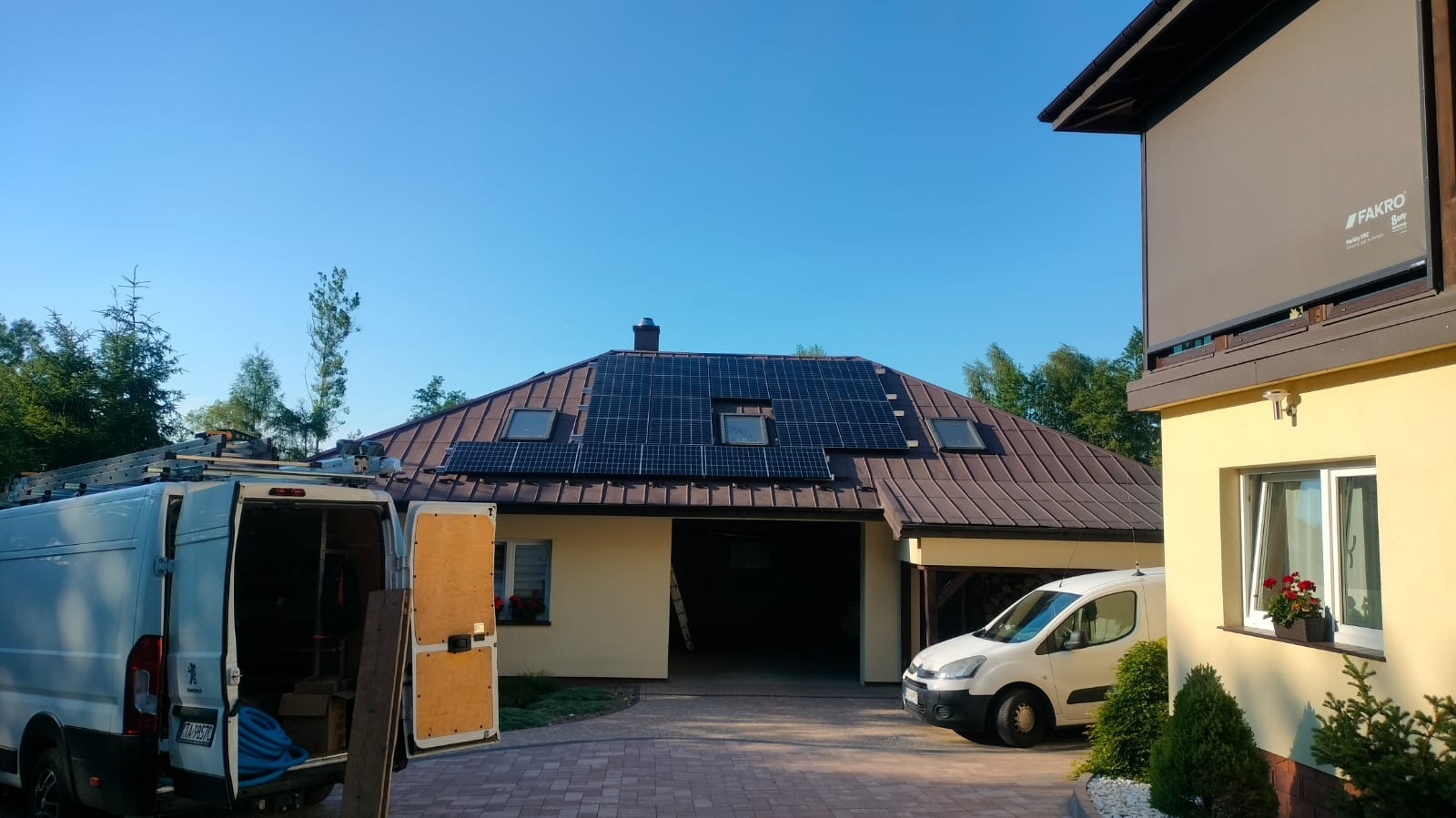 Dom jednorodzinny z brązowym dachem, na którym zamontowano panele fotowoltaiczne między oknami dachowymi, na podjeździe stoją dwa busy, jeden z otwartymi drzwiami, w tle drzewa i błękitne niebo.