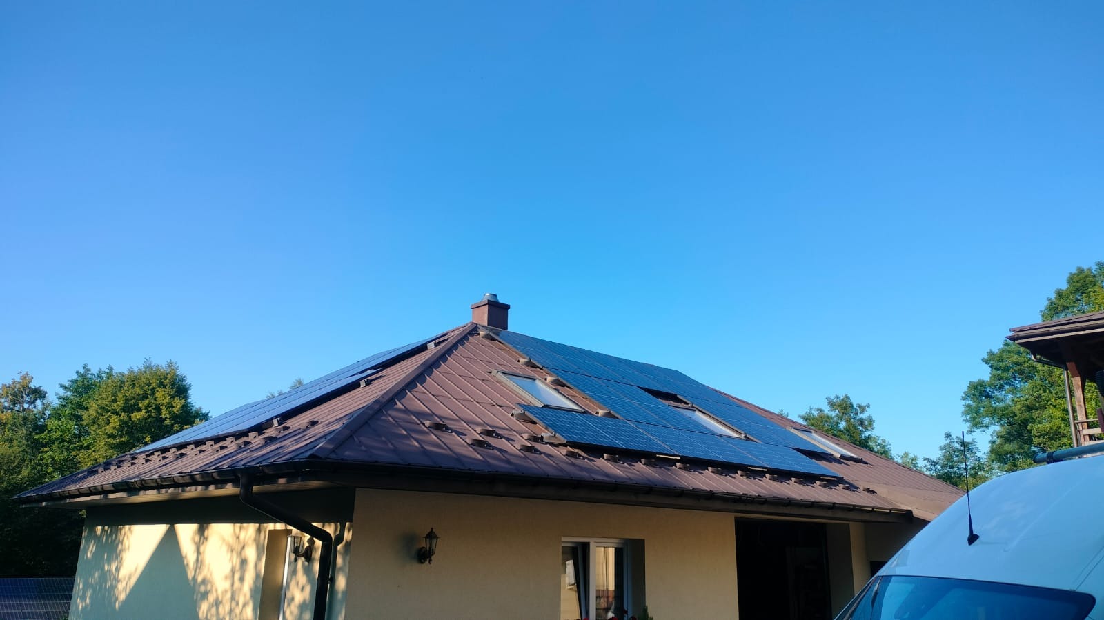 Brązowy dach z panelami słonecznymi i oknami dachowymi na tle błękitnego nieba. Częściowo widoczny biały samochód dostawczy z anteną.