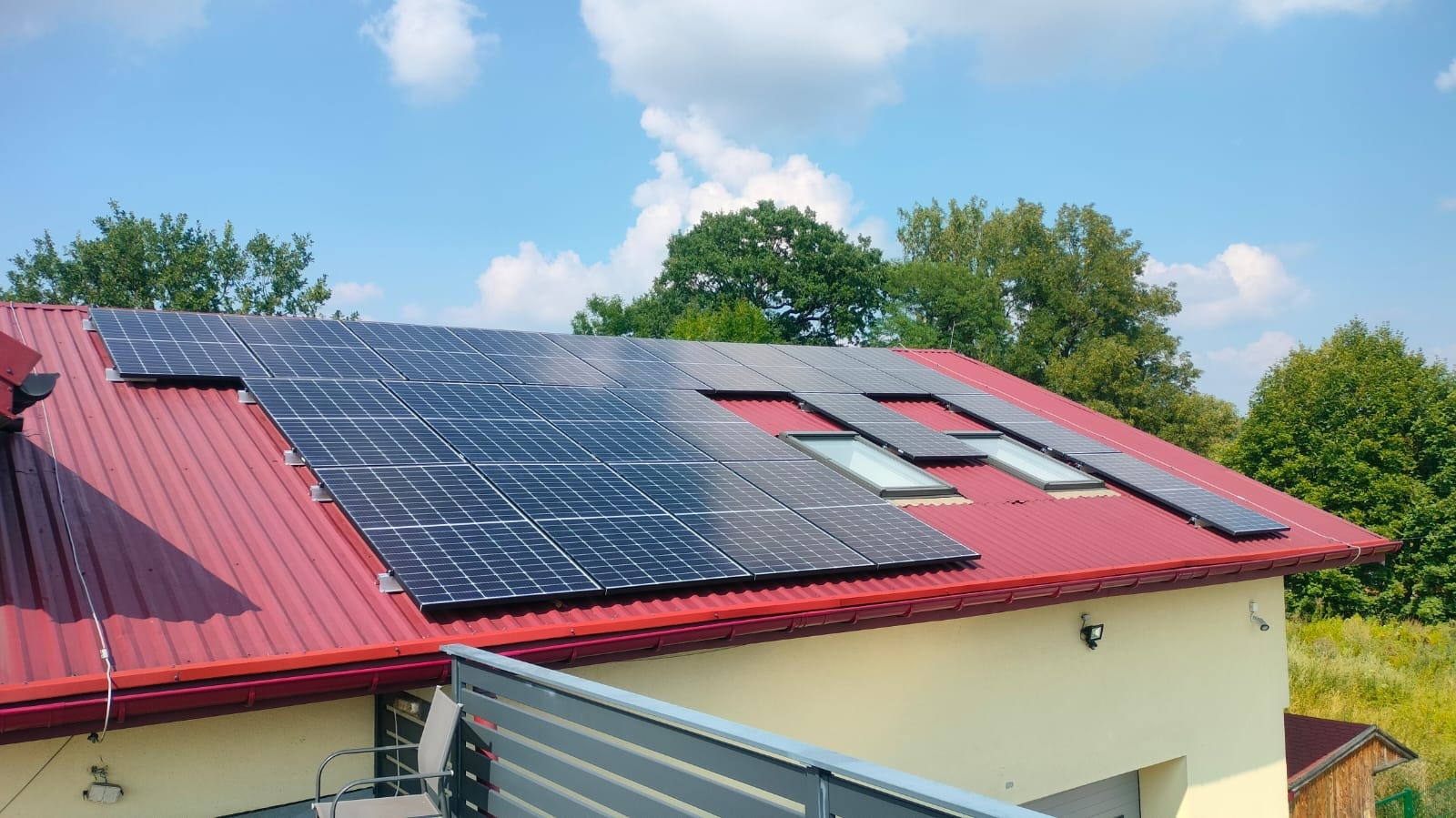 Czerwony blaszany dach z panelami słonecznymi i oknami dachowymi na tle zieleni i błękitnego nieba.