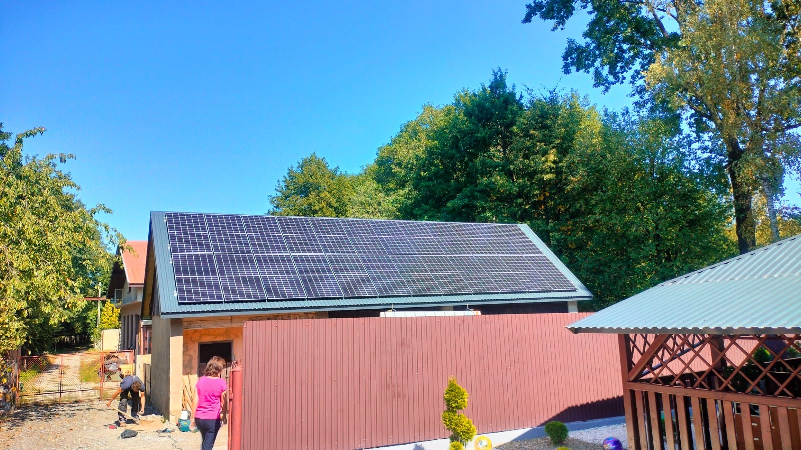 Gotowe panele słoneczne na dachu budynku gospodarczego, widoczne z poziomu gruntu, na tle zieleni i błękitnego nieba; na pierwszym planie brązowe ogrodzenie z blachy trapezowej i fragment altany.