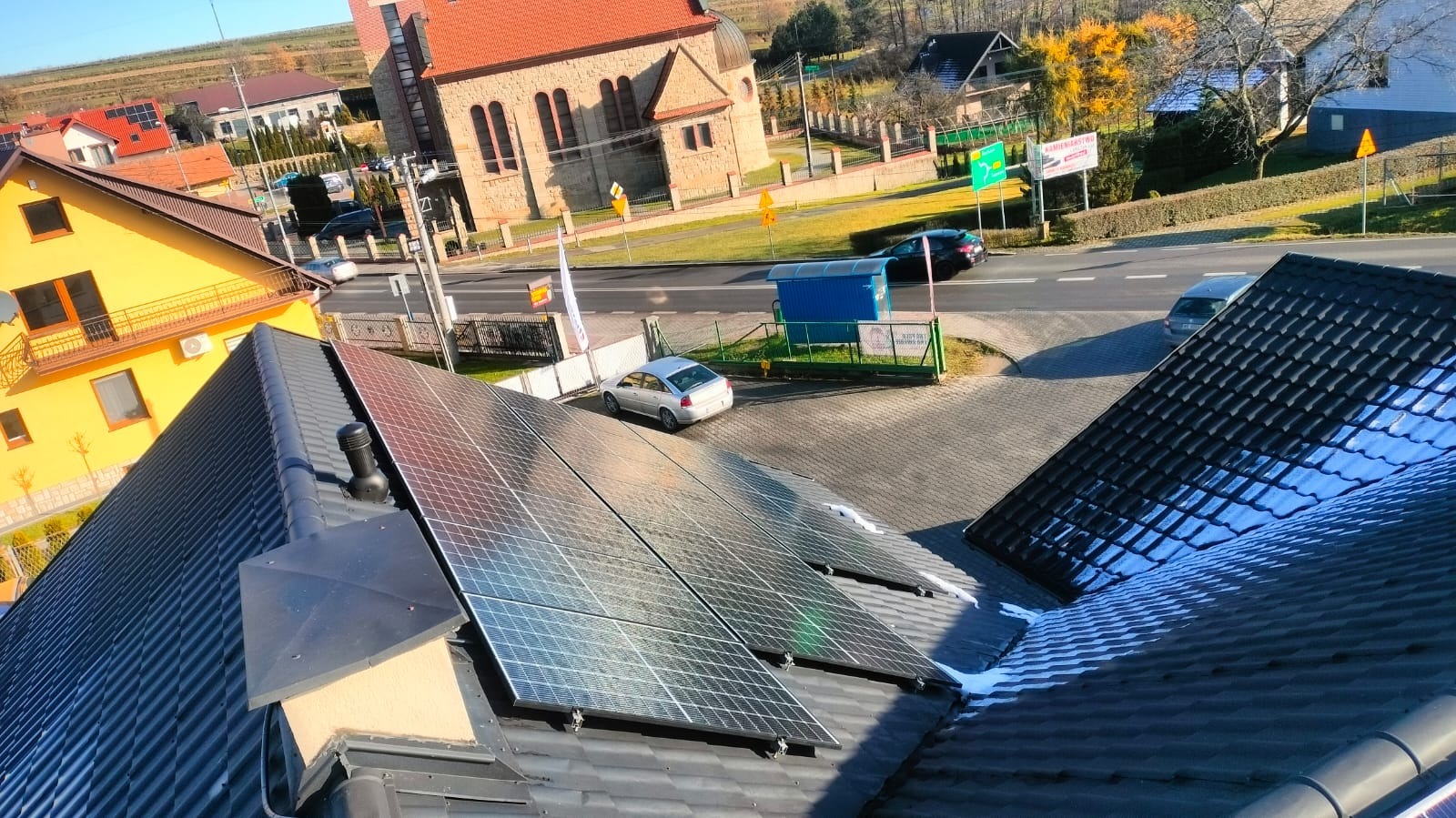 Panel słoneczny na dachu domu z widokiem na ulicę, kościół i inne budynki w słoneczny dzień.