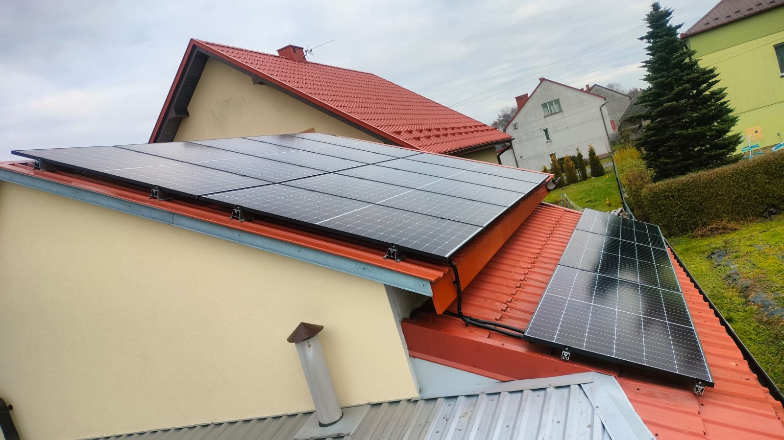 Panele słoneczne zamontowane na czerwonym, blaszanym dachu domu jednorodzinnego z kremową elewacją, widoczne uchwyty mocujące, w tle zabudowa mieszkalna i zieleń.