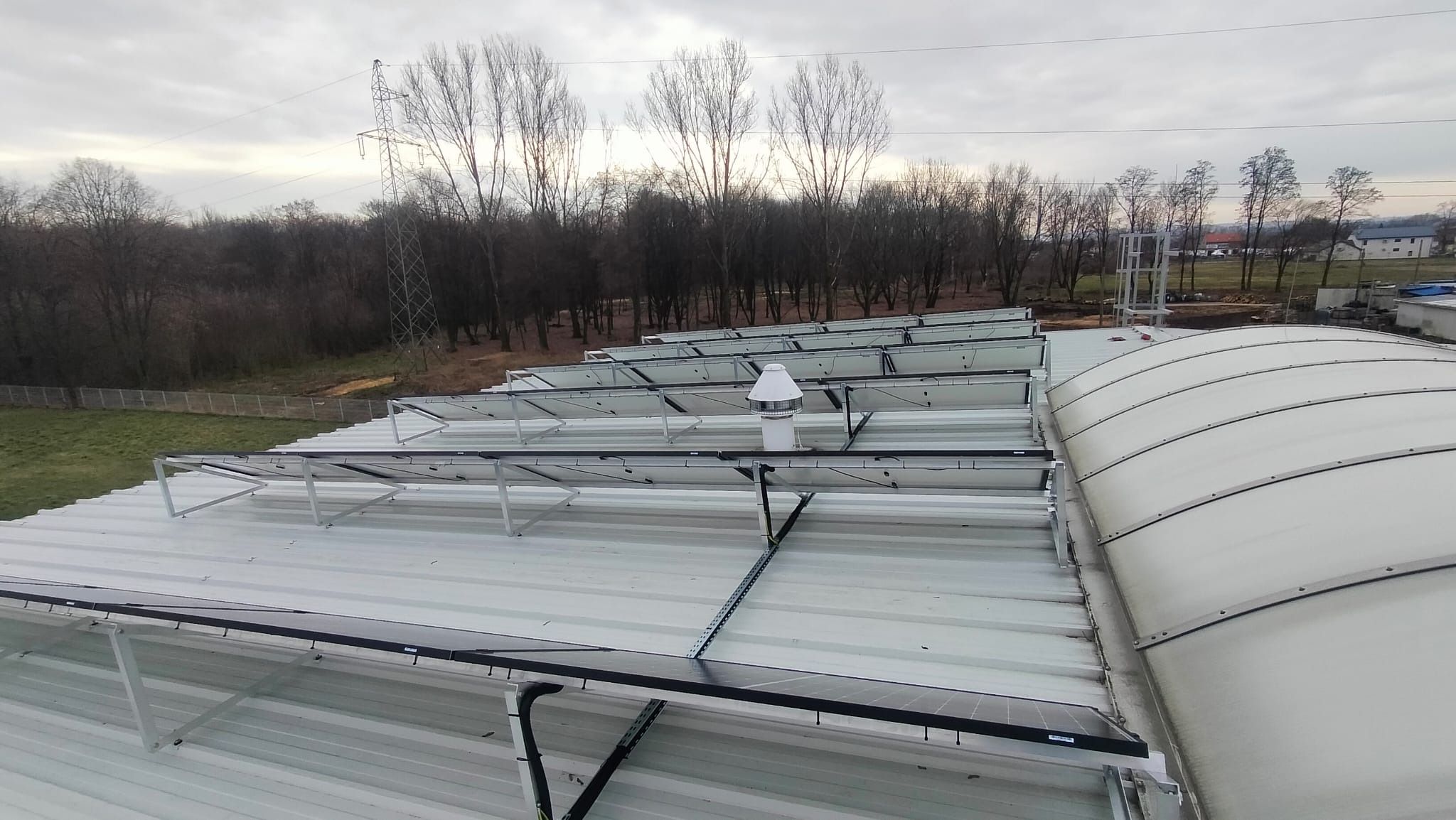 Rząd paneli słonecznych na falistym dachu z blachy trapezowej, z kominkiem wentylacyjnym w tle drzewa i słup wysokiego napięcia.