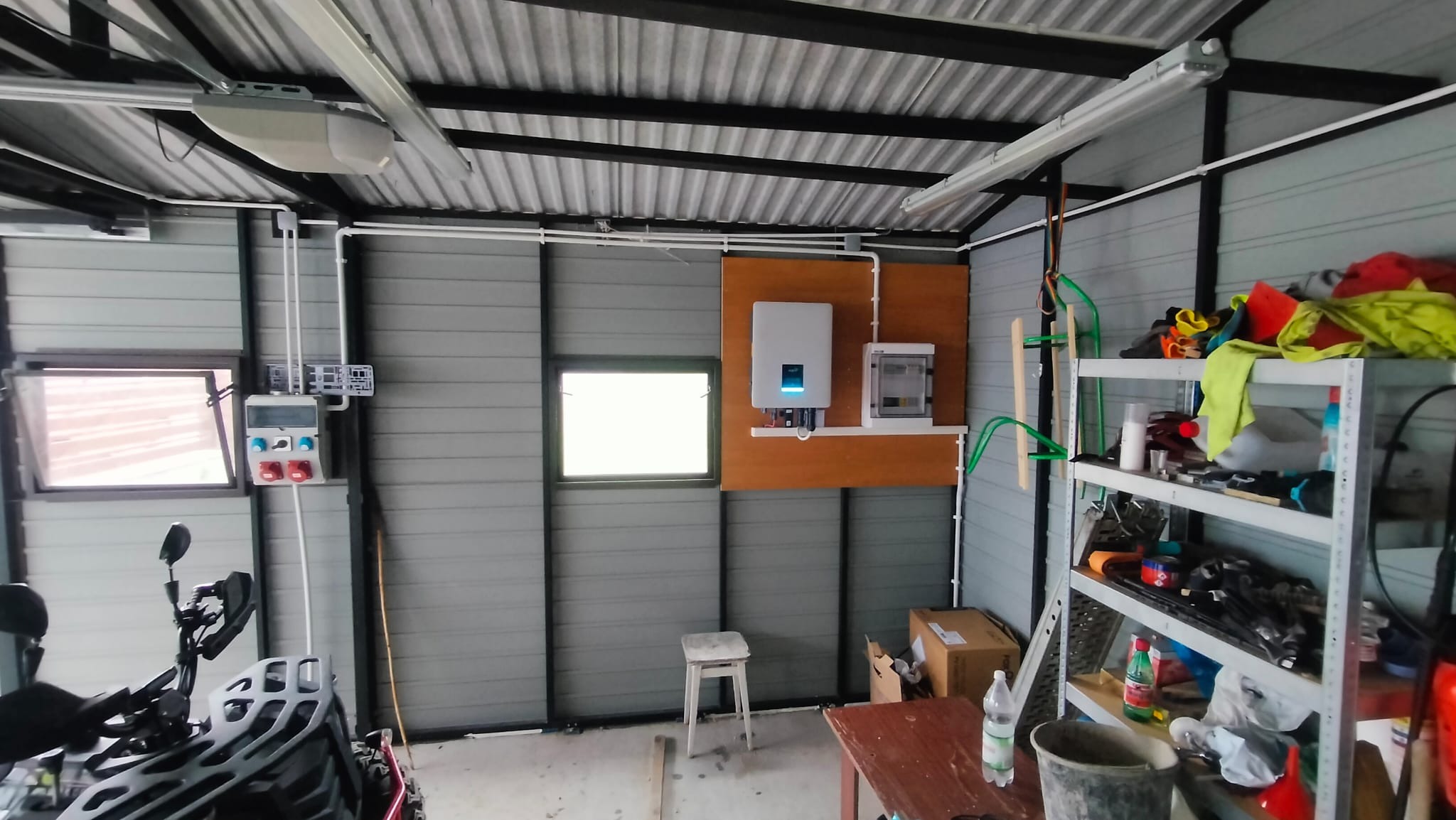 Wnętrze garażu z zamontowanym inwerterem fotowoltaicznym i skrzynką elektryczną na drewnianej płycie, widoczny motocykl crossowy, regał z narzędziami i akcesoriami.