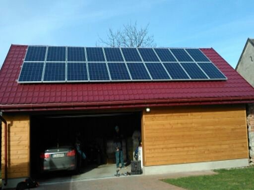 Panele słoneczne na dachu garażu z czerwoną dachówką, częściowo otwarte wrota garażowe z widocznym samochodem w środku oraz dwoma osobami w tle, błękitne niebo