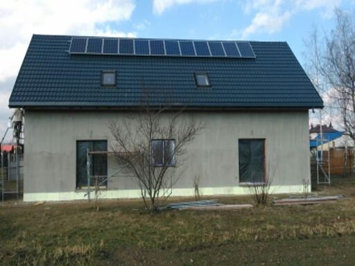 Dom jednorodzinny z szarą elewacją i ciemnym dachem z panelami słonecznymi, widoczne rusztowania przy ścianie bocznej, okna zabezpieczone folią, w tle zabudowa sąsiedzka.