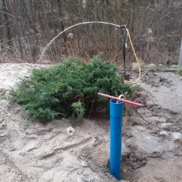Testowanie wydajności nowej studni: Woda tryska z rury, zasilana pompą, na tle piaszczystego terenu z krzewem i fragmentem ogrodu.
