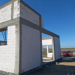 Surowy stan domu z białego bloczka, widoczny szkielet konstrukcji, betonowe nadproża, drabina w oknie, czerwony samochód z otwartymi drzwiami i osoba w tle.