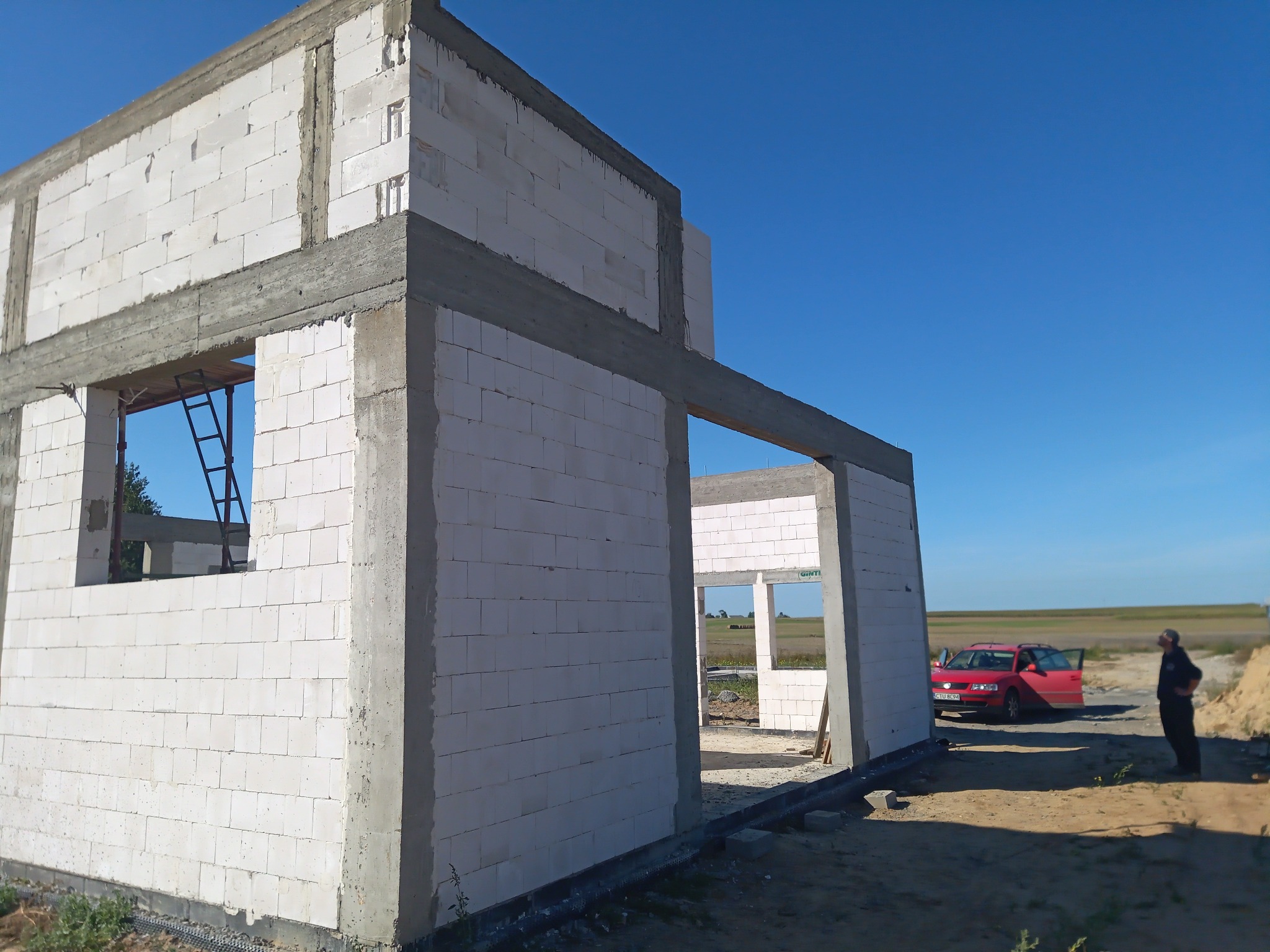 Surowy stan domu z białego bloczka, widoczny szkielet konstrukcji, betonowe nadproża, drabina w oknie, czerwony samochód z otwartymi drzwiami i osoba w tle.