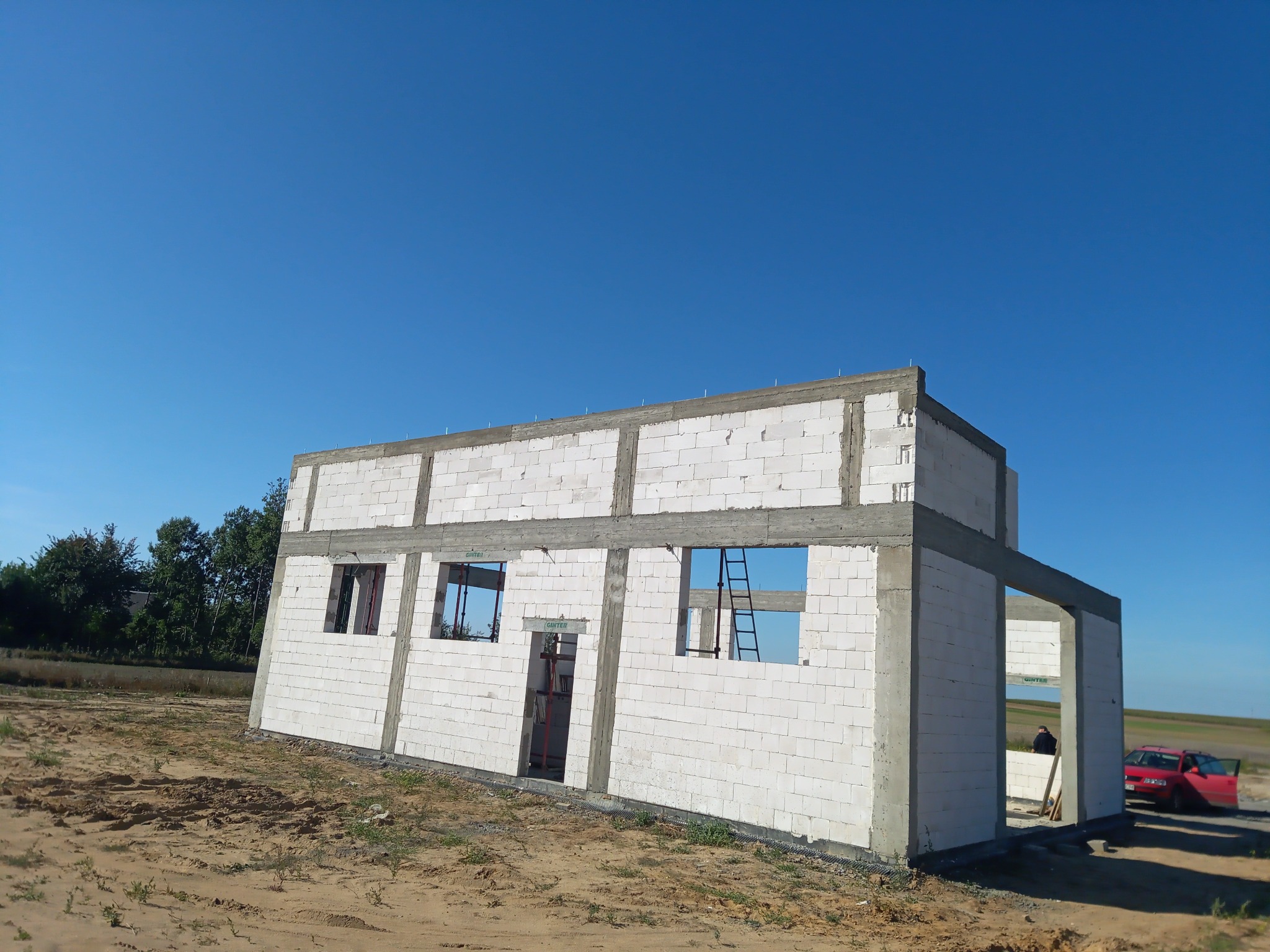 Surowy stan domu z białych bloczków, widoczne otwory okienne i drzwiowe, szare nadproża, teren budowy z piaskiem i czerwony samochód w tle.