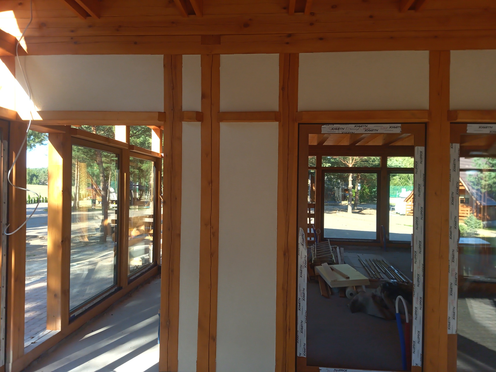 Wnętrze domu w trakcie budowy: drewniana konstrukcja szkieletowa z widocznymi oknami i elementami izolacji, w tle widok na zewnątrz.