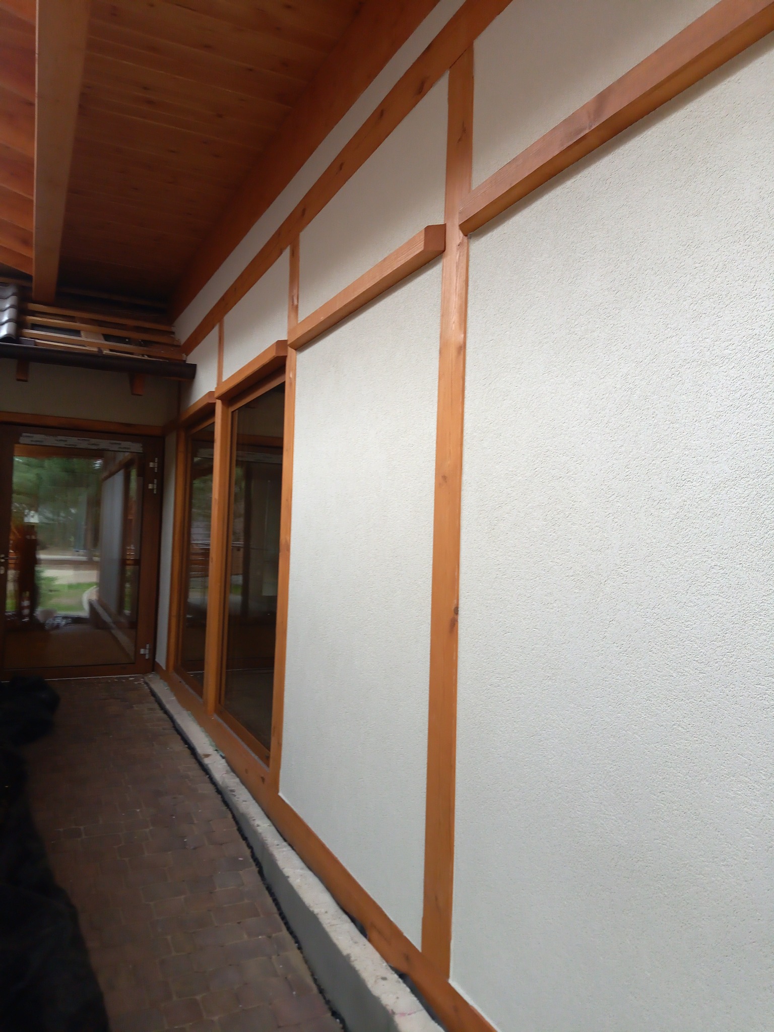 Elewacja domu z jasnym tynkiem i drewnianą konstrukcją szkieletową, widoczne okna i ceglana ścieżka.