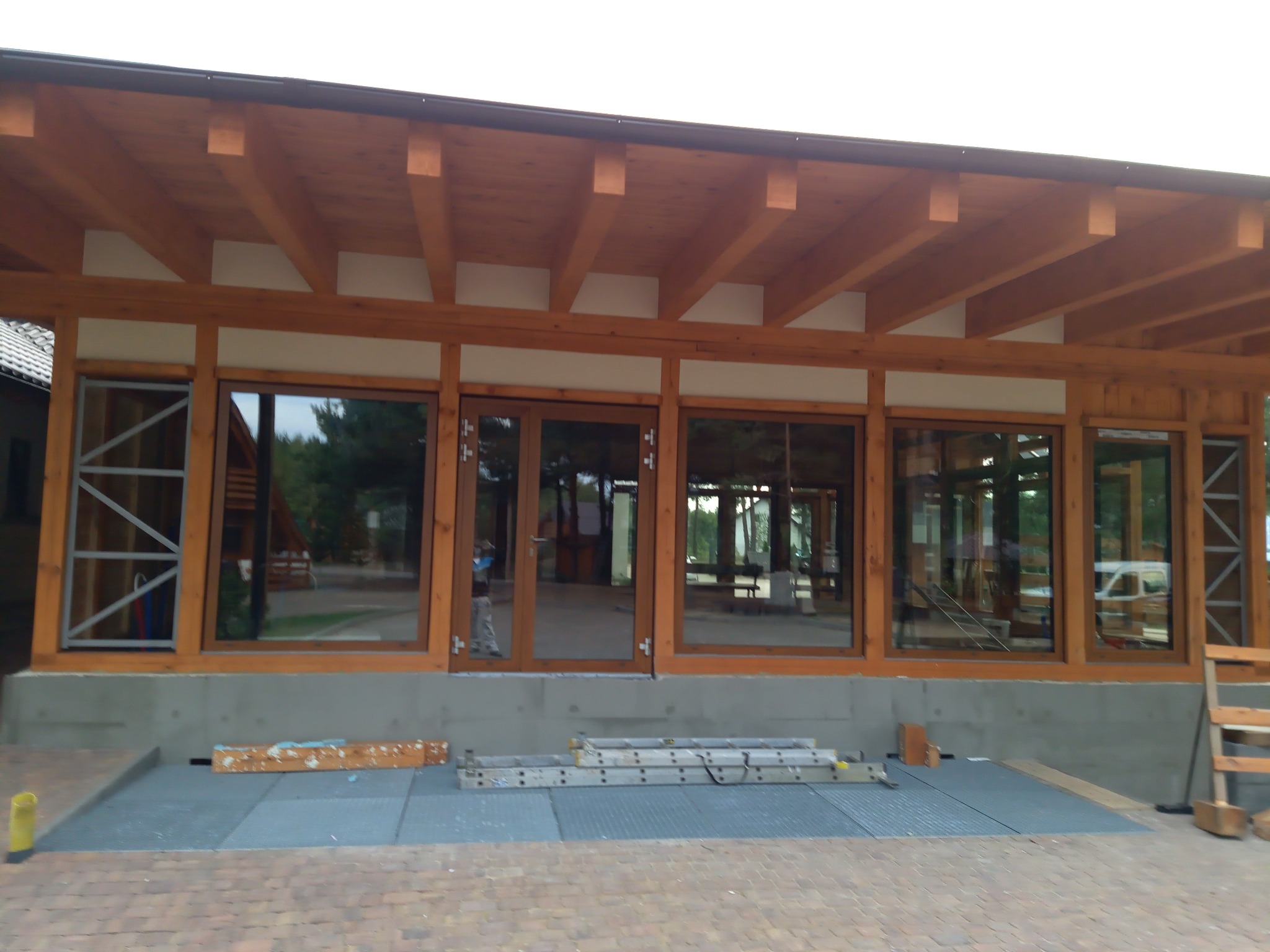 Budynek w trakcie budowy z drewnianą konstrukcją dachu, widoczne okna i drzwi z drewnianymi ramami, drabiny i materiały budowlane na zewnątrz.