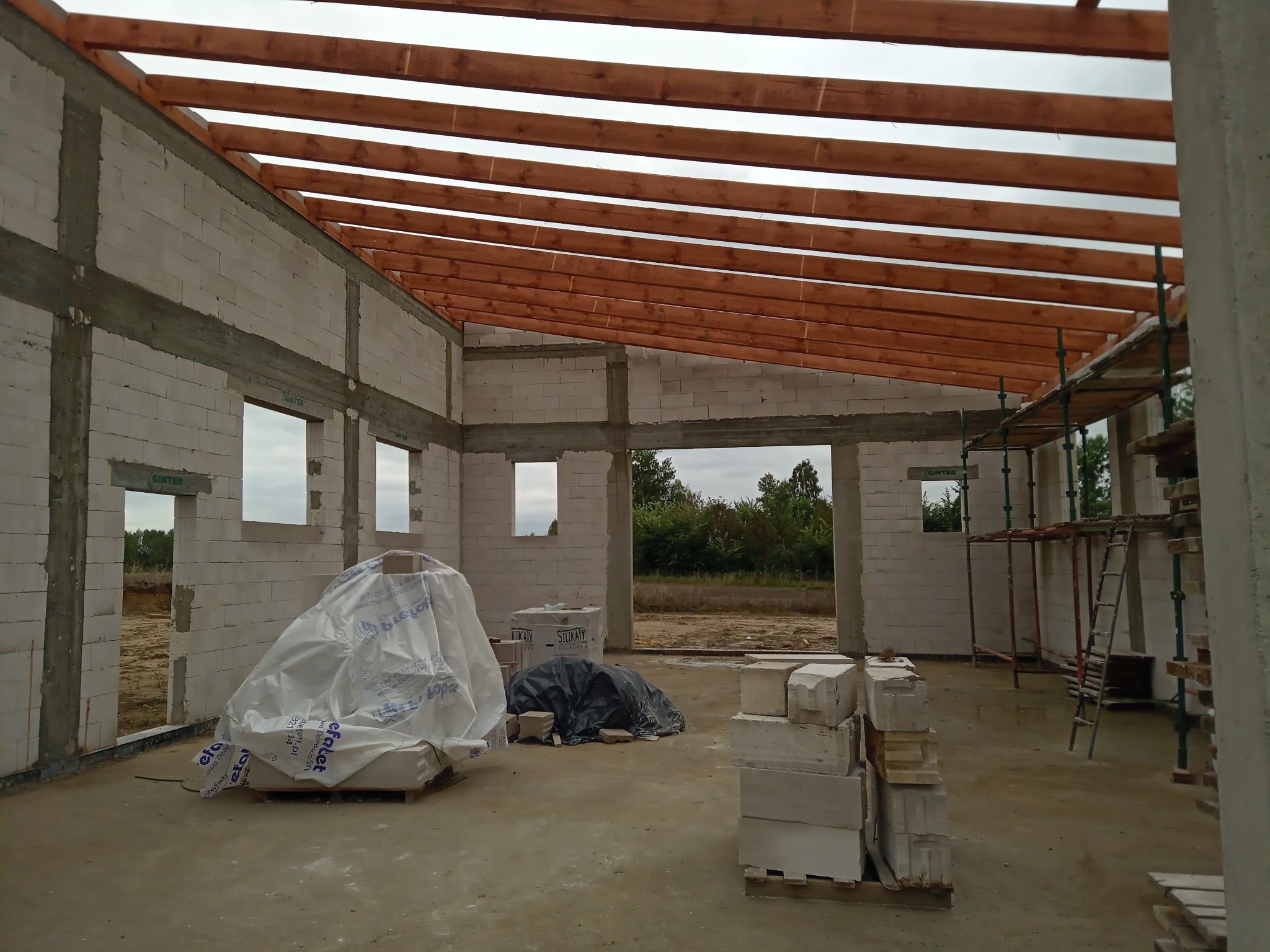 Wnętrze surowego domu w trakcie budowy z widocznymi ścianami z betonu komórkowego, drewnianą konstrukcją dachu, rusztowaniami i materiałami budowlanymi, przykrytymi folią ochronną.