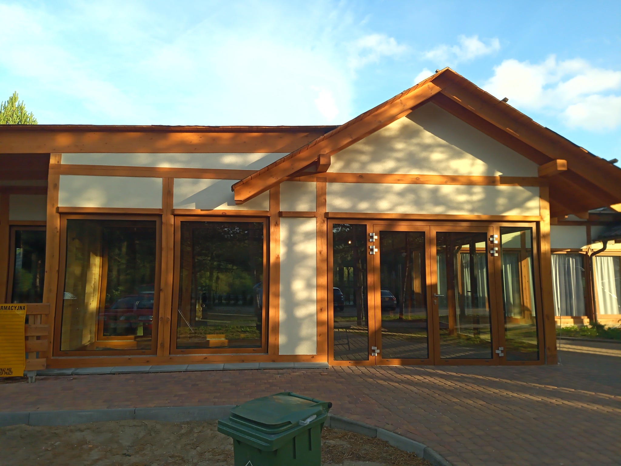 Budynek parterowy z drewnianą konstrukcją szkieletową i dużymi oknami, widoczny kosz na śmieci z przodu, ceglana ścieżka prowadząca do wejścia.