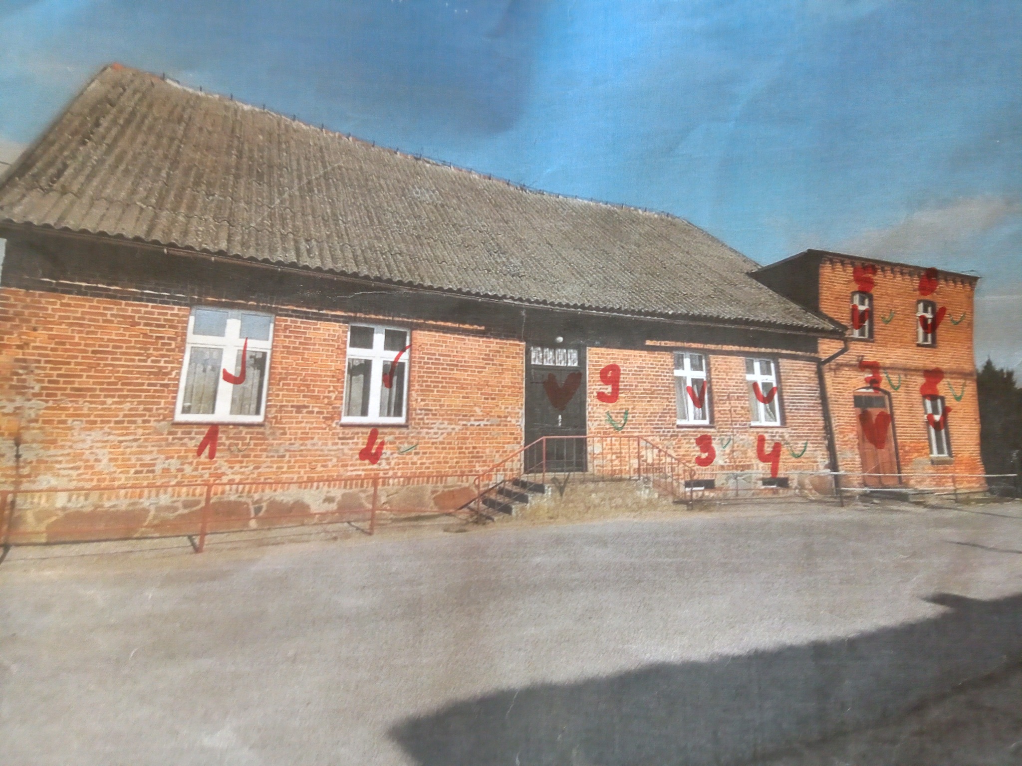 Dwukondygnacyjny budynek murowany z cegły z szarym dachem krytym eternitem, widoczne okna z białymi ramami, metalowa balustrada przed budynkiem, ślady graffiti na elewacji.