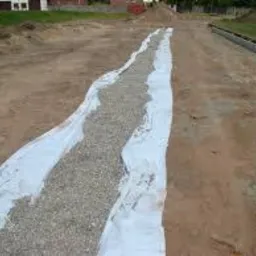 Drenaż liniowy w trakcie budowy: biała geowłóknina wyścielająca wykop wypełniony żwirem na tle ziemi i trawnika.