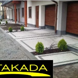 Jasnoszara kostka brukowa z ciemnymi obramowaniami tworzy wzór przed domem z brązowymi bramami garażowymi i rabatami z tujami. Uwagę przyciąga napis ESTAKADA.