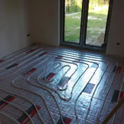 Układanie ogrzewania podłogowego w nowym domu. Białe rurki na folii z czerwoną siatką, okno w tle. Widoczne niedoskonałości ścian w surowym stanie.